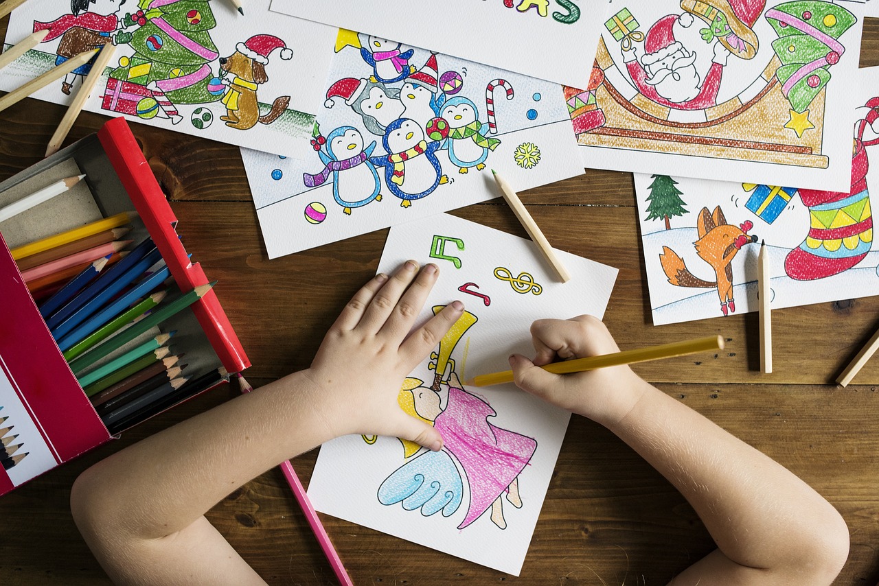 background image of a child's arms colouring a picture of a christmas angel playing a trumpet surrounded by other coloured pictures and a box of colourful pencil crayons